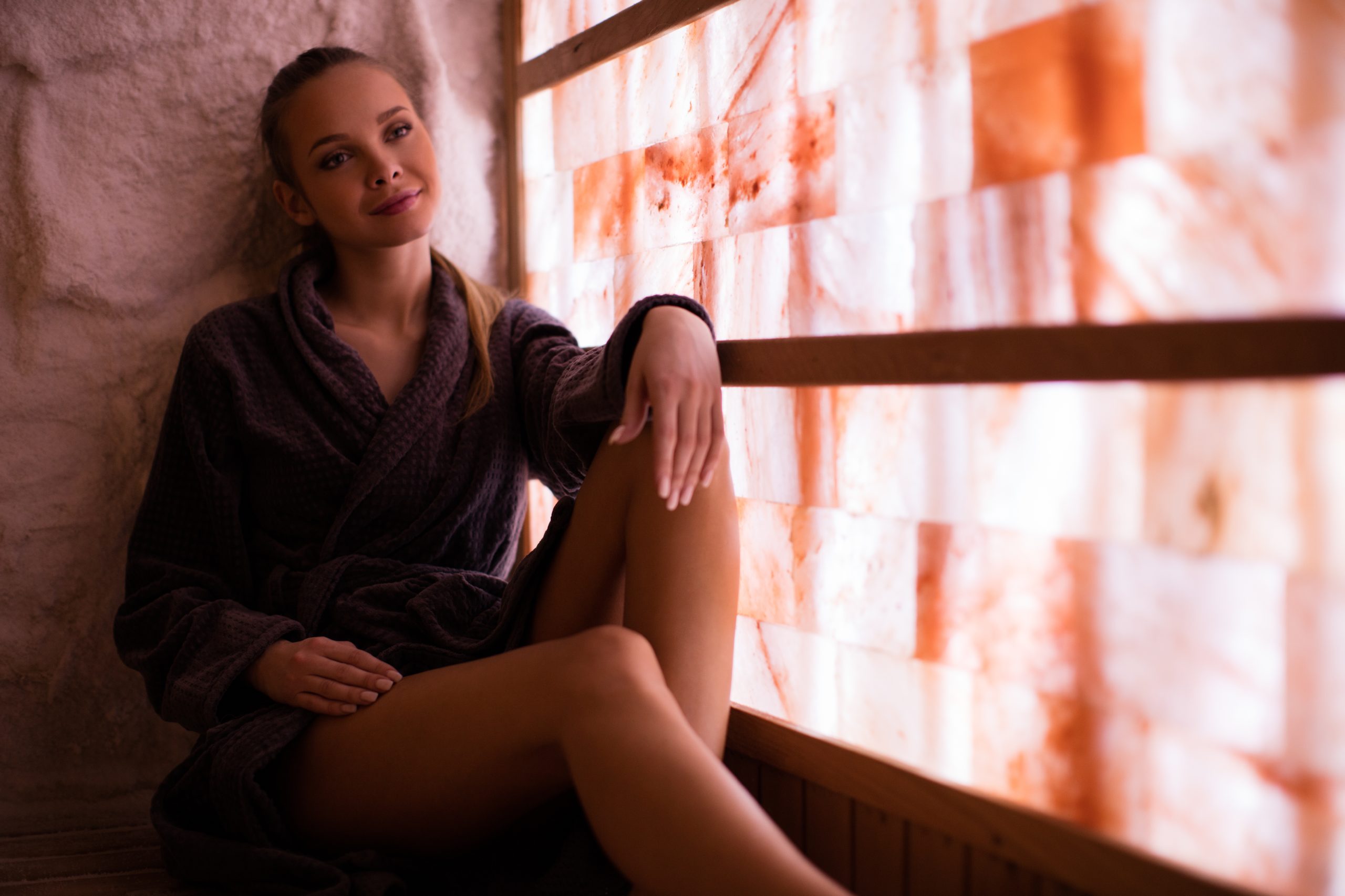 Beautiful young woman relaxing in salt room and enjoying in halotherapy treatment. She wearing bathrobe and sitting on the wooden bench and in front of her is transparent glowing wall made of salt crystals. Photo taken under the available light from glowing wall.